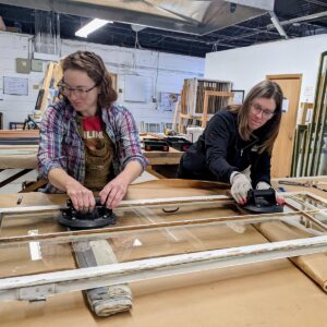 Two women repairing a window frame in a workshop.